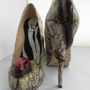 Könnte beinhalten: Ein Paar High-Heel-Schuhe mit Schlangenhautmuster. Die Absätze sind schlangenförmig mit einem roten Kopf. Die Schuhe sind mit "Calle" beschriftet.