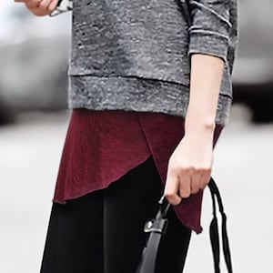 Op de afbeelding: Een vrouw draagt een grijs sweatshirt, een bordeauxrode rok, zwarte leggings en zwart-witte sneakers. Ze draagt een zwarte quilted tote bag met gouden accenten.