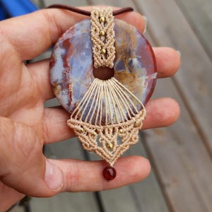 May include: A circular pendant featuring a polished, multi-colored stone with swirls of red, brown, and white. The stone is encased in beige macrame cord with small gold-toned beads and a single dark red bead hanging from the bottom center.