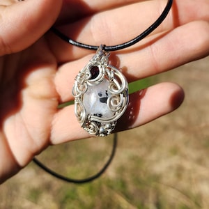 May include: A wire-wrapped pendant featuring a central oval white stone with black inclusions and a small dark red accent stone. The silver-toned metal wire is coiled in intricate loops around the stones, suspended on a black cord necklace.