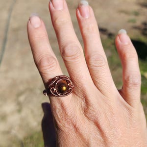 May include: A wire-wrapped ring featuring a central round brown bead with a metallic gold sheen. The band is constructed from thin, coiled copper-colored wire that forms an intricate, layered pattern around the circular stone setting.