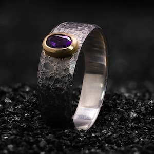 May include: A silver ring with a textured band and a gold setting. The setting holds an oval, deep purple gemstone. The ring is displayed on a dark, textured surface, highlighting its metallic sheen and gemstone color.