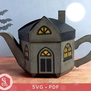 Peut inclure: Une théière artisanale en papier sur le thème d'Halloween en forme de maison hantée. La maison grise a un toit noir, des fenêtres éclairées en jaune et une cheminée. Trois découpes de chauves-souris volent au-dessus du bec. Le design comprend le texte "SVG • PDF".