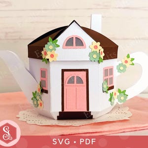 May include: A white paper teapot shaped like a house, with a brown roof and pink door and windows. The teapot is decorated with paper flowers and sits on a pink cloth. The image includes the text "SVG • PDF".