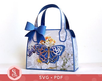Könnte beinhalten: Eine blaue und weiße Papiertüte mit einem Schmetterlingsdesign und einer blauen Schleife. Die Tasche hat einen Henkel und ist perfekt zum Verschenken.