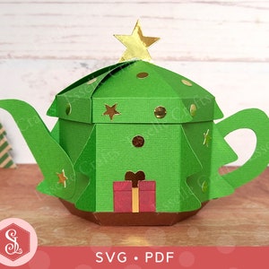 Puede incluir: Una tetera de papel verde con forma de árbol de Navidad con una estrella dorada en la parte superior. La tetera tiene una base marrón y detalles dorados. La tetera está sobre una superficie de madera. SVG. PDF