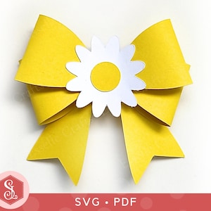 Pode incluir: Um laço de papel amarelo brilhante com uma margarida branca no centro. A margarida tem um centro amarelo. O laço tem pontas afiadas. A imagem também inclui o texto "SVG • PDF" e um logotipo.