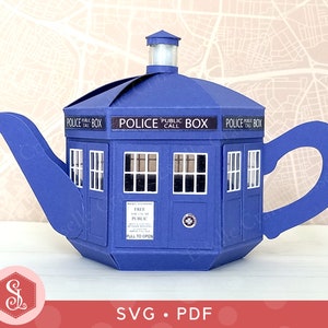 Può includere: Una teiera di carta blu a forma di cabina telefonica della polizia britannica. La teiera ha un beccuccio, un manico e un coperchio con una luce. Le parole "POLICE PUBLIC BOX" sono stampate sulla parte superiore. Il design include finestre e una porta.