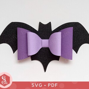 May include: A black felt bat shape with a purple bow in the center. The bat has pointed wings and a notched head. The bow is made of paper and has a textured appearance. The bottom of the image has the text "SVG • PDF".
