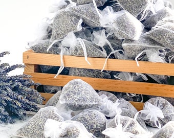 May include: A wooden crate filled with small white mesh bags filled with dried lavender. The bags are tied with white string and are spilling out of the crate onto a white surface.