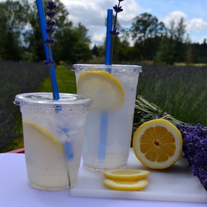 May include: Two clear plastic cups filled with lemonade, garnished with lemon slices and lavender sprigs. Blue straws are inserted into the cups. The cups are sitting on a white cutting board, which is on top of a red metal surface.