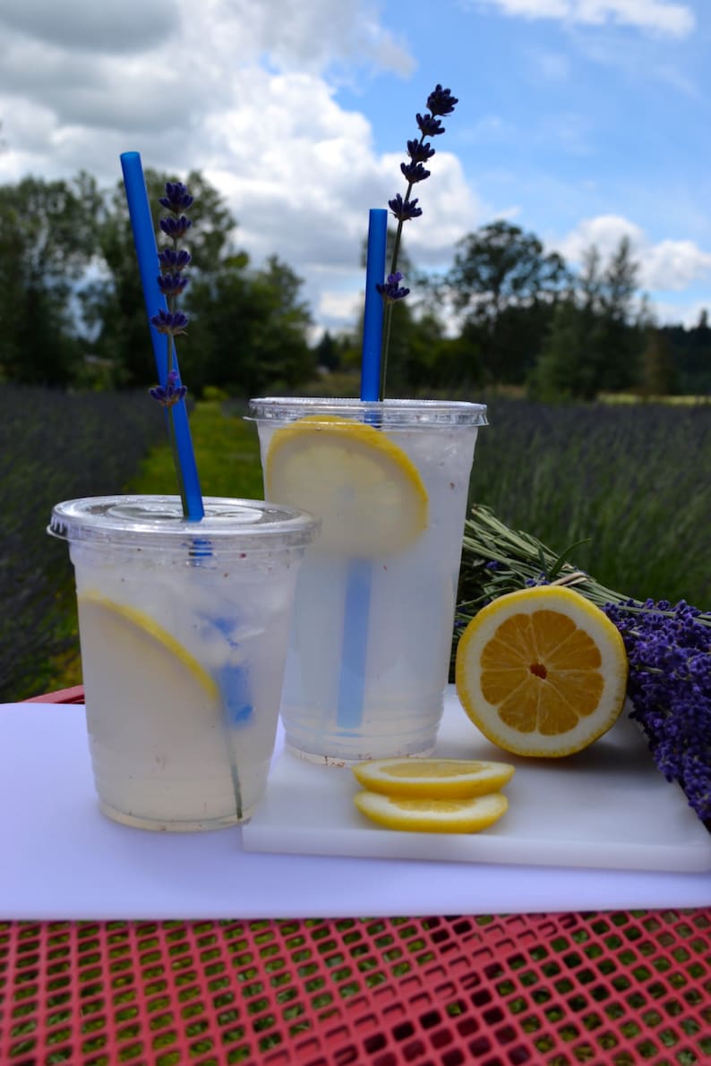 May include: Two clear plastic cups filled with lemonade, garnished with lemon slices and lavender sprigs. Blue straws are inserted into the cups. The cups are sitting on a white cutting board, which is on top of a red metal surface.