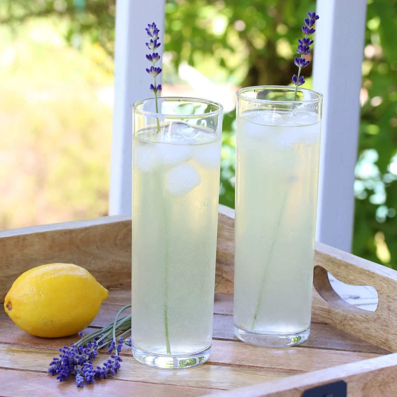 May include: Two tall glasses of lemonade with ice cubes and lavender sprigs. The glasses are on a wooden tray with a lemon beside them.
