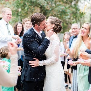 Puede incluir: Una pareja de reci&eacute;n casados se besa, rodeada de confeti y de invitados alegres. La novia lleva un vestido de novia de encaje blanco, y el novio un traje oscuro. La imagen captura una alegre celebraci&oacute;n de boda.