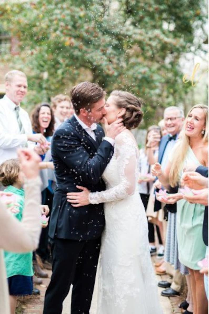 Puede incluir: Una pareja de reci&eacute;n casados se besa, rodeada de confeti y de invitados alegres. La novia lleva un vestido de novia de encaje blanco, y el novio un traje oscuro. La imagen captura una alegre celebraci&oacute;n de boda.