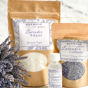 May include: Three brown paper bags with white labels containing lavender sugar, culinary lavender, and lavender extract. The labels feature the text "Norwood Farm" and the product name. The bags are sitting on a gold wire rack. A bunch of lavender flowers is in the foreground.