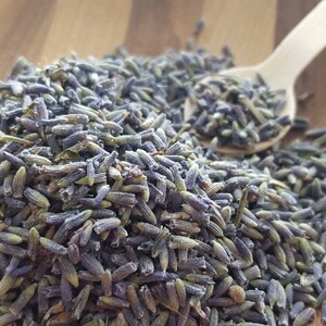 Puede incluir: Primer plano de capullos de lavanda secos, de color morado claro, sobre una superficie de madera. Una peque&ntilde;a cuchara de madera est&aacute; presente, lo que sugiere un uso culinario o de aromaterapia.