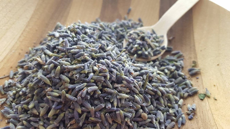 Puede incluir: Primer plano de capullos de lavanda secos, de color morado claro, sobre una superficie de madera. Una peque&ntilde;a cuchara de madera est&aacute; presente, lo que sugiere un uso culinario o de aromaterapia.