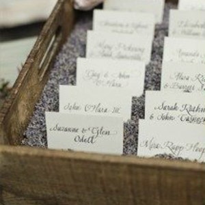Puede incluir: Una bandeja de madera llena de lavanda y tarjetas de sitio. Las tarjetas son blancas con nombres de caligraf&iacute;a elegante. La bandeja es r&uacute;stica y la lavanda a&ntilde;ade un elemento decorativo fragante, adecuado para una boda o evento.
