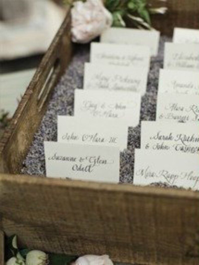 Puede incluir: Una bandeja de madera llena de lavanda y tarjetas de sitio. Las tarjetas son blancas con nombres de caligraf&iacute;a elegante. La bandeja es r&uacute;stica y la lavanda a&ntilde;ade un elemento decorativo fragante, adecuado para una boda o evento.