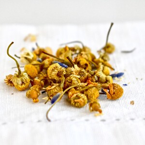 May include: A close-up of dried chamomile flowers and lavender buds on a white linen surface. The flowers are a pale yellow color and the buds are a light blue.