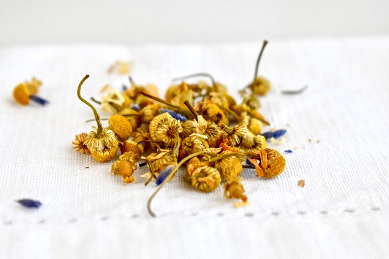 May include: A close-up of dried chamomile flowers and lavender buds on a white linen surface. The flowers are a pale yellow color and the buds are a light blue.