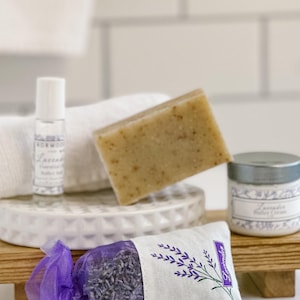 May include: A lavender-themed spa gift set with a small bottle of lavender bath oil, a bar of lavender soap, a jar of lavender bath cream, and a lavender-scented sachet. The items are arranged on a white ceramic tray and a wooden platform.