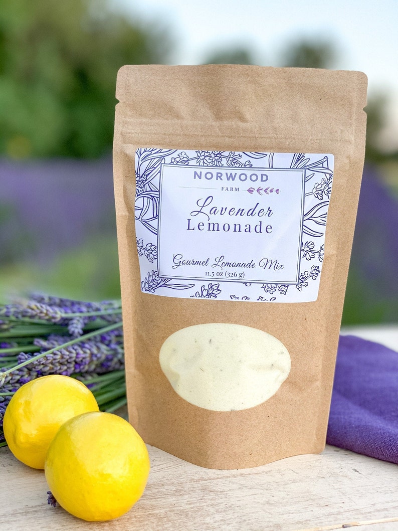 May include: A brown paper bag with a white label that says "Norwood Farm Lavender Lemonade Gourmet Lemonade Mix 11.5 oz (326 g)". The label has a floral design with lavender flowers. There are two lemons and a bunch of lavender flowers in the background.