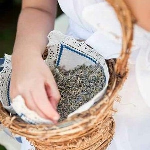 Puede incluir: Una cesta redonda de mimbre llena de capullos de lavanda secos, forrada con un pa&ntilde;o blanco ribeteado con encaje y costuras azules. La cesta tiene un asa curva. Una persona lleva un vestido blanco.