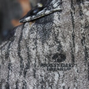 May include: Close-up of a camouflage pattern fabric, featuring shades of gray, brown, and black, with the text "Mossy Oak Break-Up" visible. The fabric appears to be part of a garment, possibly a shirt or jacket.