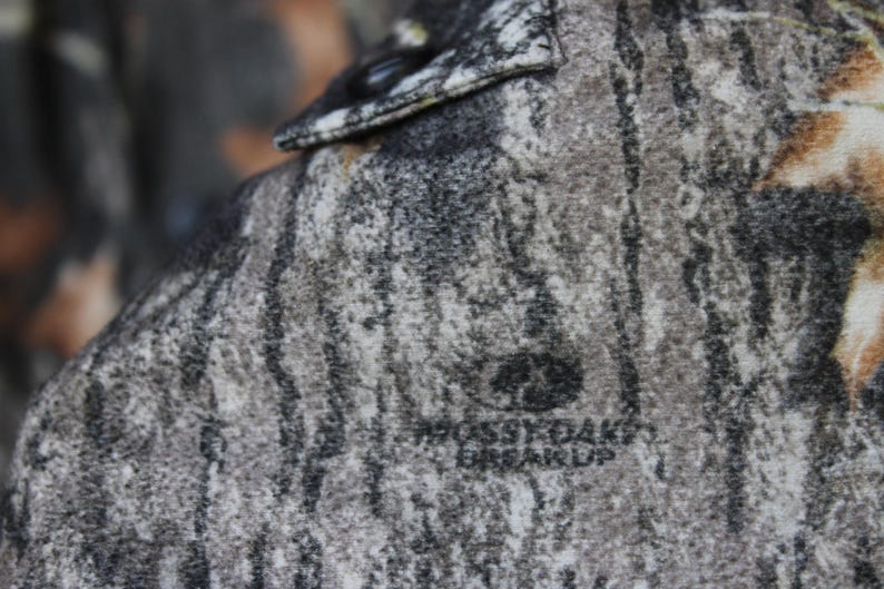 May include: Close-up of a camouflage pattern fabric, featuring shades of gray, brown, and black, with the text "Mossy Oak Break-Up" visible. The fabric appears to be part of a garment, possibly a shirt or jacket.