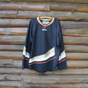 May include: Black hockey jersey with gold, white, and orange accents. The jersey has long sleeves and a v-neck collar with a Reebok logo. The jersey is hanging on a wooden hanger against a wooden background.
