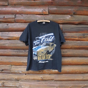 Könnte beinhalten: Schwarzes T-Shirt mit einer Grafik eines Oldtimers in Blau und Gelb, mit dem Text "Too Fast To Rust" und "Snap-on". Das T-Shirt hängt an einem Holzbügel vor einem Holzgrund.