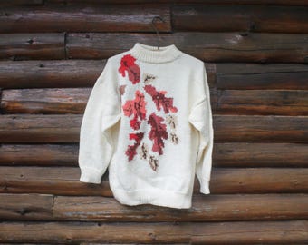 May include: White knitted sweater with a colorful leaf design. The sweater has a high neck and long sleeves.