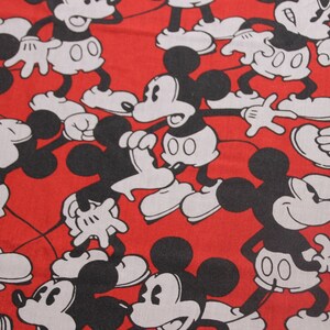Op de afbeelding: Rode stof met een herhalend patroon van zwarte en witte Mickey Mouse-figuren. De figuren staan in verschillende poses, waaronder zwaaien, glimlachen en er verrast uitzien.