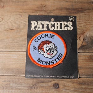 Könnte beinhalten: Vintage "Cookie Monster" Aufnäher auf einer schwarzen Trägerkarte. Der Aufnäher zeigt ein Cartoon-Bild des Krümelmonsters mit orangefarbenem Haar und einem weißen Gesicht, umgeben von einem orangefarbenen Rand. Auf der Karte steht "PATCHES" in einer Vintage-Schrift.