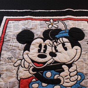 Puede incluir: Manta acolchada con Mickey y Minnie Mouse abraz&aacute;ndose. El dise&ntilde;o incluye un fondo de impresi&oacute;n de peri&oacute;dico, un borde rojo y un borde exterior en blanco y negro. Minnie lleva un sombrero azul con una flor.