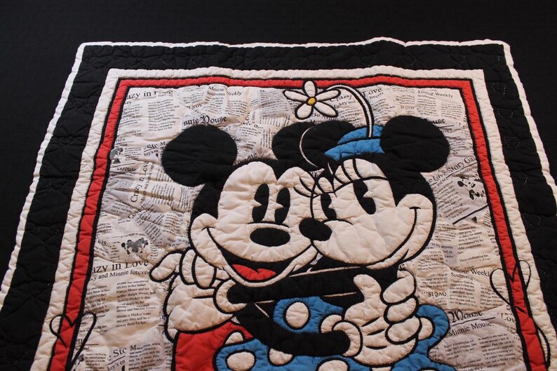 Puede incluir: Manta acolchada con Mickey y Minnie Mouse abraz&aacute;ndose. El dise&ntilde;o incluye un fondo de impresi&oacute;n de peri&oacute;dico, un borde rojo y un borde exterior en blanco y negro. Minnie lleva un sombrero azul con una flor.