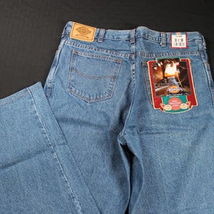 Puede incluir: Un par de pantalones vaqueros azules de mezclilla con un parche de cuero marrón en la parte posterior de la cintura. Los pantalones tienen una pierna recta y un diseño clásico de cinco bolsillos. El bolsillo trasero tiene una etiqueta roja y blanca con el texto "Dickies Workwear" y una imagen de un tren.