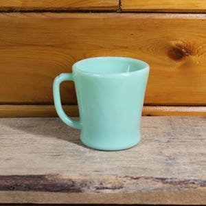 Può includere: Una tazza vintage verde chiaro con un manico curvo. La tazza ha una base arrotondata e un bordo leggermente svasato. La tazza è realizzata in un materiale solido e opaco e poggia su una superficie di legno.
