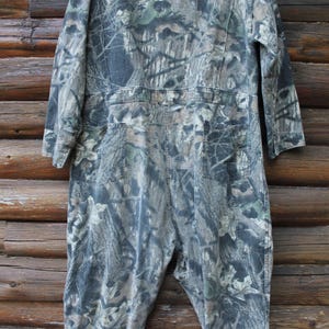 May include: A camouflage jumpsuit in shades of green, brown, and grey. The one-piece garment features long sleeves, a collared neckline, and multiple pockets. The jumpsuit is made of a durable fabric, suitable for outdoor activities.