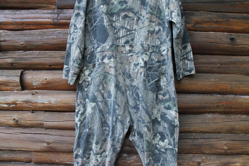 May include: A camouflage jumpsuit in shades of green, brown, and grey. The one-piece garment features long sleeves, a collared neckline, and multiple pockets. The jumpsuit is made of a durable fabric, suitable for outdoor activities.