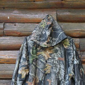 May include: Camouflage hooded jacket with a button-up front. The jacket features a pattern of green, brown, and tan leaves and branches. The jacket is hanging against a wooden background.