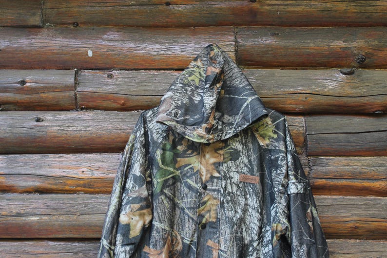 May include: Camouflage hooded jacket with a button-up front. The jacket features a pattern of green, brown, and tan leaves and branches. The jacket is hanging against a wooden background.