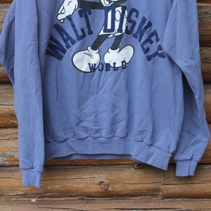 Puede incluir: Sudadera azul con un gr&aacute;fico de Mickey Mouse y la inscripci&oacute;n "WALT DISNEY WORLD" en azul marino. Tiene mangas largas y pu&ntilde;os y dobladillo acanalados. El fondo es una superficie de madera.