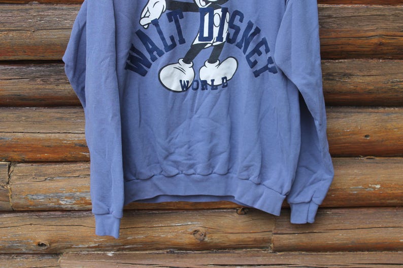 Puede incluir: Sudadera azul con un gr&aacute;fico de Mickey Mouse y la inscripci&oacute;n "WALT DISNEY WORLD" en azul marino. Tiene mangas largas y pu&ntilde;os y dobladillo acanalados. El fondo es una superficie de madera.