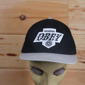 Peut inclure: Casquette de baseball noire et grise avec un logo "WorldWide OBEY" blanc sur le devant.