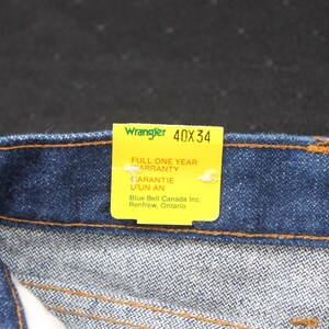 May include: A blue denim jean waistband with a yellow tag that reads "Wrangler 40X34" and "Full One Year Warranty" in English and French. The tag also includes the company name and location: "Blue Bell Canada Inc. Renfrew, Ontario."