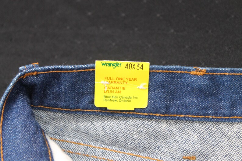 May include: A blue denim jean waistband with a yellow tag that reads "Wrangler 40X34" and "Full One Year Warranty" in English and French. The tag also includes the company name and location: "Blue Bell Canada Inc. Renfrew, Ontario."