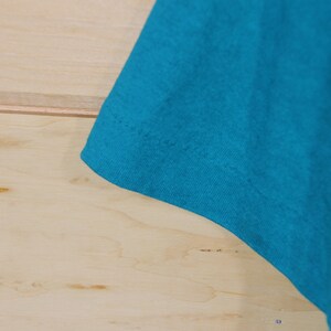 May include: Close-up of the turquoise blue fabric of a t-shirt. The fabric is smooth and has a slight sheen.
